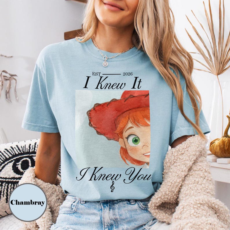 May include: A light blue short-sleeve t-shirt featuring a watercolor illustration of a character with red hair and a red hat. The text reads EST 2026 I Knew It I Knew You with a treble clef symbol. The shirt has a crew neckline and a relaxed fit.