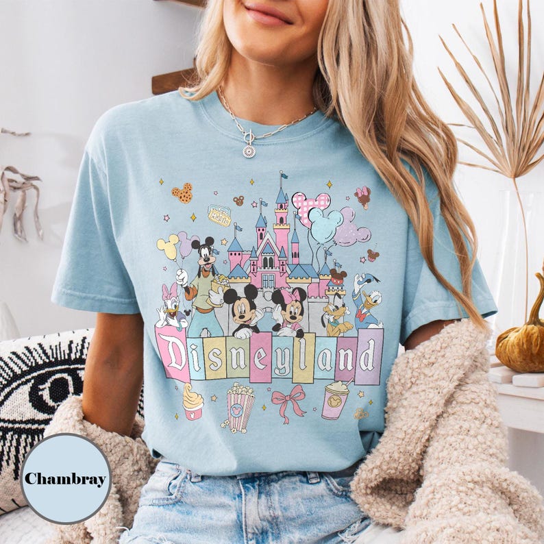 May include: Light blue t-shirt featuring a Disneyland graphic. The design includes a castle, Mickey and Minnie Mouse, Donald Duck, and other characters. The word "Disneyland" is written in pastel colors. The shirt is made of a soft material.