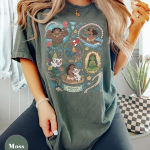 May include: Moss green t-shirt featuring a colorful graphic of characters from the animated film "Moana." The design includes illustrations of Maui, Moana, Hei Hei, Pua, and other elements. The words "Moana & Friends" are also visible.