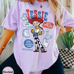 May include: A lilac-colored t-shirt featuring a graphic of the character Jessie from the "Toy Story" franchise. The design includes the character's name, a speech bubble, and the phrase "Deluxe Cowgirl Doll." The shirt also has the text "From The Hit Television Show!"