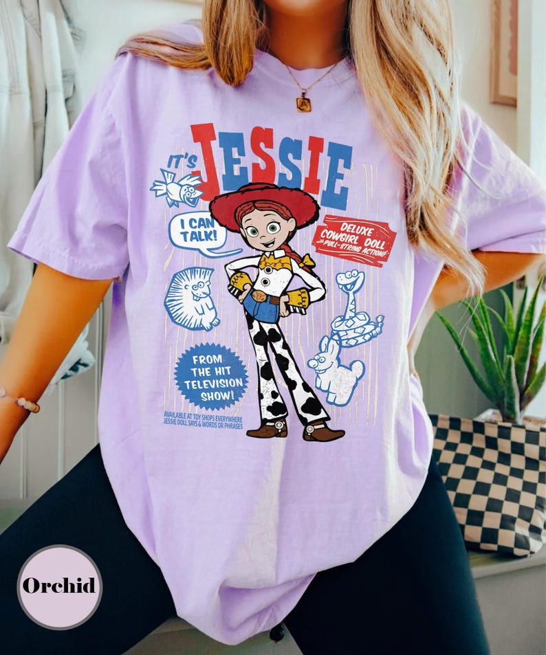 May include: A lilac-colored t-shirt featuring a graphic of the character Jessie from the "Toy Story" franchise. The design includes the character's name, a speech bubble, and the phrase "Deluxe Cowgirl Doll." The shirt also has the text "From The Hit Television Show!"
