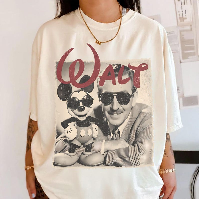 May include: Cream-colored t-shirt featuring a vintage-style graphic of Mickey Mouse and Walt Disney wearing sunglasses. The word "WALT" is printed in a burgundy script above the image. The shirt has short sleeves and a relaxed fit.