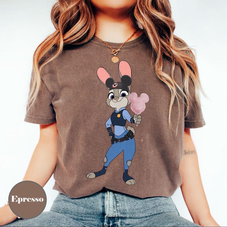 Puede incluir: Camiseta marr&oacute;n con una imagen de dibujos animados de Judy Hopps de Zootopia, vestida con uniforme de polic&iacute;a y sosteniendo un algod&oacute;n de az&uacute;car rosa. La camiseta tiene mangas cortas y cuello redondo.
