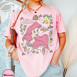 Puede incluir: Camiseta rosa claro con un dise&ntilde;o de Ariel de La Sirenita de Disney. El dise&ntilde;o muestra a Ariel con pelo rosa, rodeada de criaturas marinas. La camiseta tiene mangas cortas y cuello redondo.