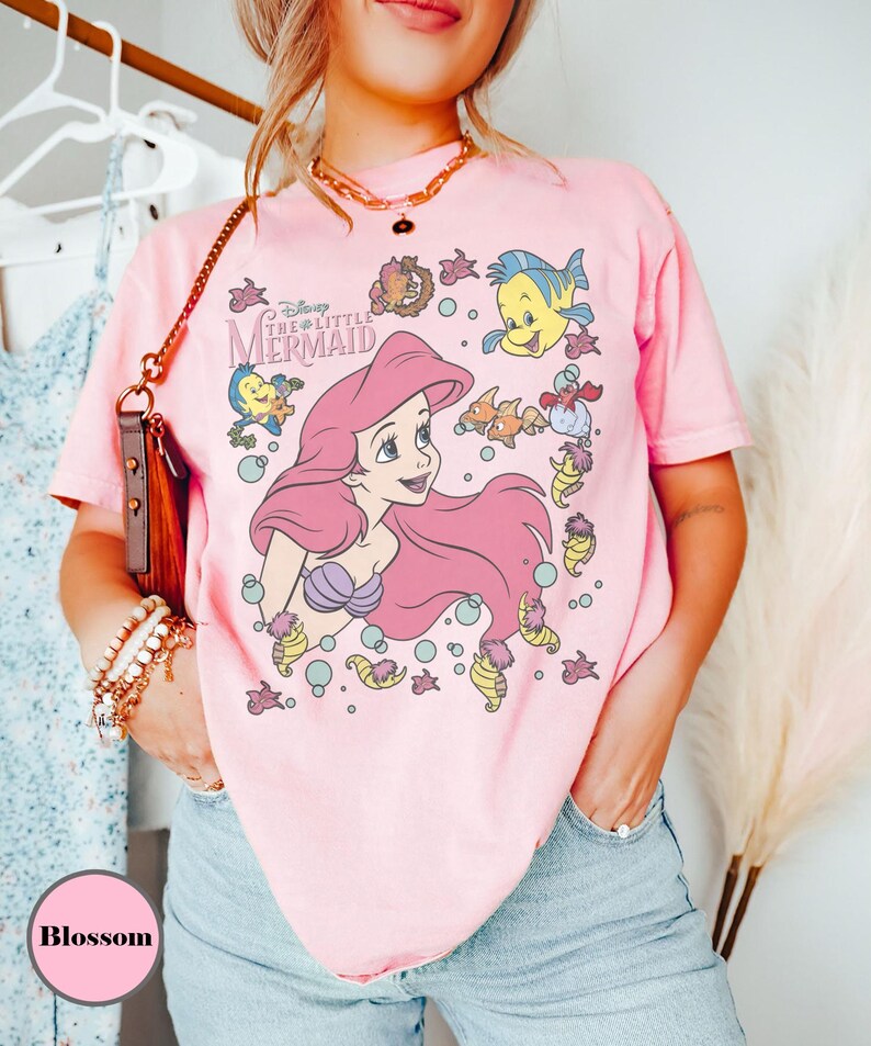 Puede incluir: Camiseta rosa claro con un dise&ntilde;o de Ariel de La Sirenita de Disney. El dise&ntilde;o muestra a Ariel con pelo rosa, rodeada de criaturas marinas. La camiseta tiene mangas cortas y cuello redondo.