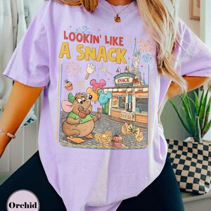 May include: Lavender t-shirt with a graphic that says "Lookin' Like A Snack." The graphic features a cartoon mouse holding balloons, with a snack stand and fireworks in the background. The shirt is a relaxed fit.