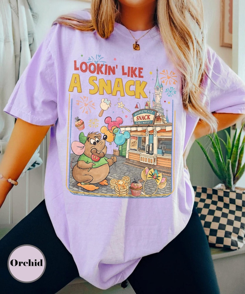 May include: Lavender t-shirt with a graphic that says "Lookin' Like A Snack." The graphic features a cartoon mouse holding balloons, with a snack stand and fireworks in the background. The shirt is a relaxed fit.