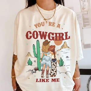 May include: An off-white short-sleeve t-shirt featuring a graphic print of two figures in cowboy hats standing in a desert landscape with cacti and mountains. The text reads YOU'RE A COWGIRL LIKE ME in bold red lettering above and below the illustration.