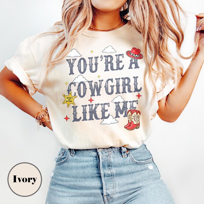 Puede incluir: Una camiseta de color marfil con el texto "YOU'RE A COWGIRL LIKE ME" en letras gris oscuro. El dise&ntilde;o incluye un sombrero de vaquero rojo, una insignia de estrella de sheriff amarilla, un par de botas vaqueras rojas, nubes blancas y estrellas rojas.