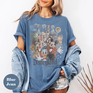 May include: Blue jean-colored t-shirt featuring a vintage Disneyland graphic. The design includes Mickey Mouse, Minnie Mouse, Donald Duck, and Goofy in front of a castle. The text "Disneyland Est 1955" is printed below the characters.