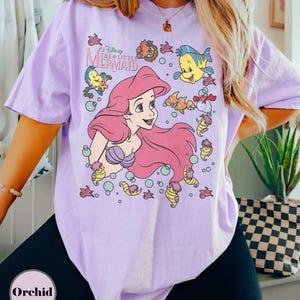 Puede incluir: Una camiseta de color lila claro con un gr&aacute;fico de Ariel de La Sirenita de Disney. El dise&ntilde;o incluye a Ariel con pelo rosa, Flounder, Sebasti&aacute;n y otras criaturas marinas. La camiseta tiene mangas cortas.