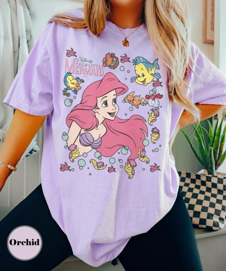 Puede incluir: Una camiseta de color lila claro con un gr&aacute;fico de Ariel de La Sirenita de Disney. El dise&ntilde;o incluye a Ariel con pelo rosa, Flounder, Sebasti&aacute;n y otras criaturas marinas. La camiseta tiene mangas cortas.