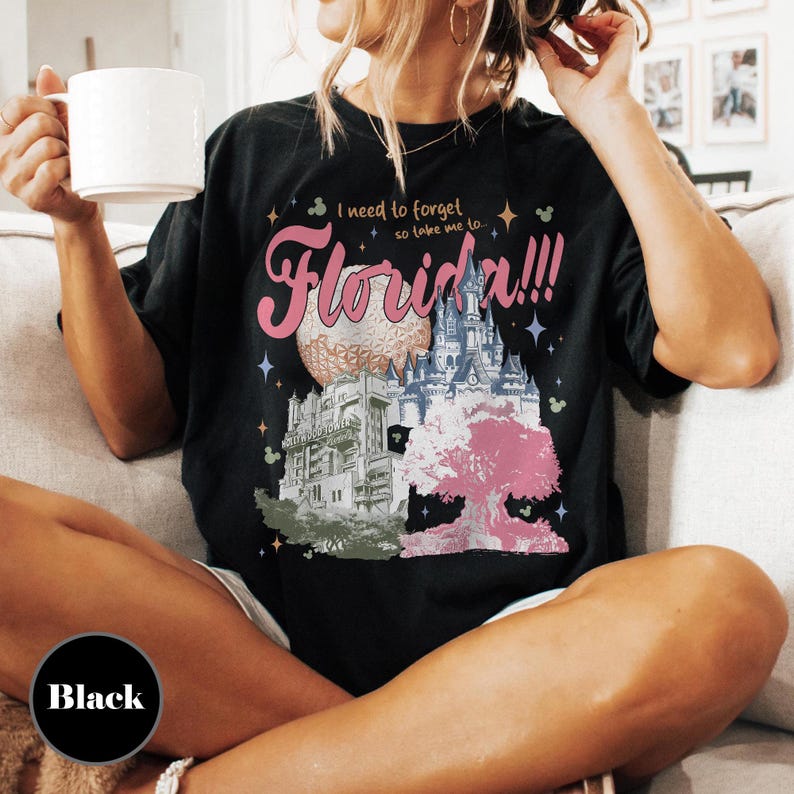 May include: Black t-shirt with a graphic design featuring the text "I need to forget so take me to... Florida!!!" The design includes illustrations of a castle, a sphere, and a pink tree. The shirt is a relaxed fit.