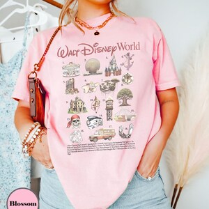 May include: A pale pink t-shirt with the words "Walt Disney World" in a cursive style. The shirt displays various Disney-themed illustrations and a list of attractions. The shirt is worn by a person.