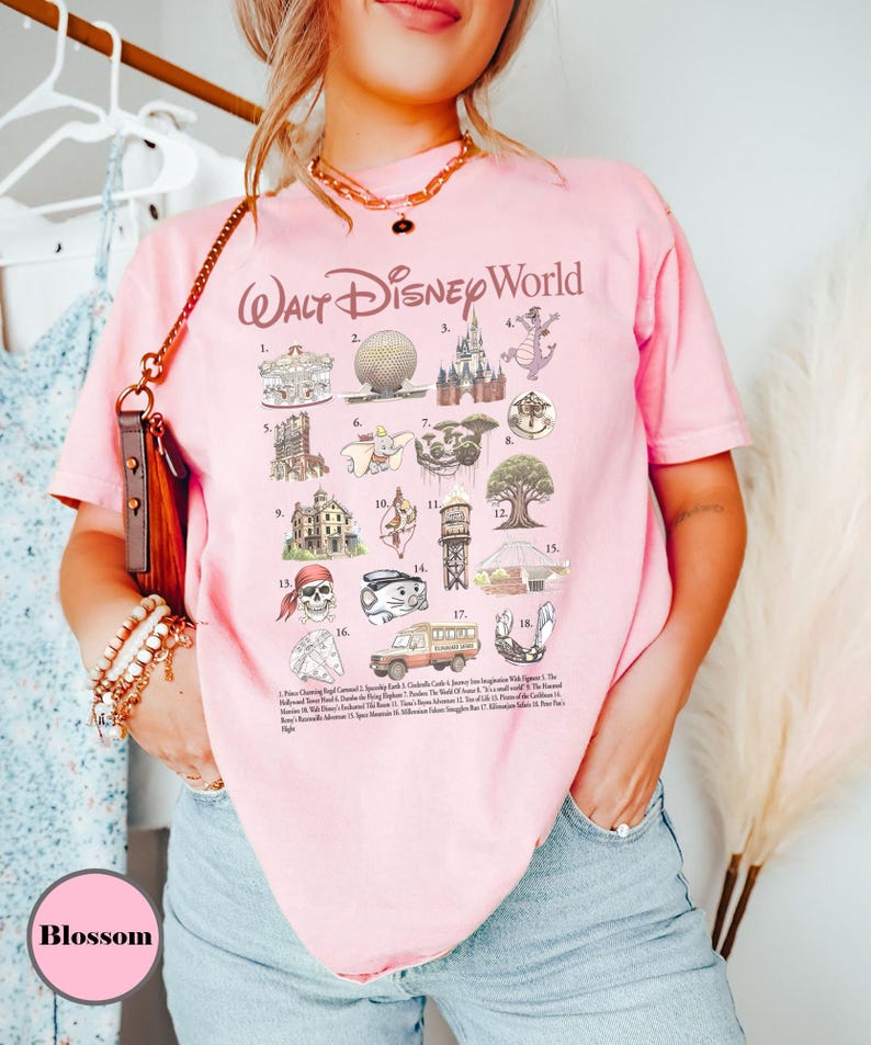 May include: A pale pink t-shirt with the words "Walt Disney World" in a cursive style. The shirt displays various Disney-themed illustrations and a list of attractions. The shirt is worn by a person.