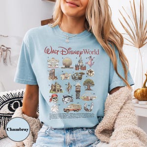 May include: Pale blue t-shirt featuring a graphic of Walt Disney World attractions. The shirt displays a carousel, Spaceship Earth, Cinderella Castle, and other landmarks. The text "Walt Disney World" is printed in burgundy script.