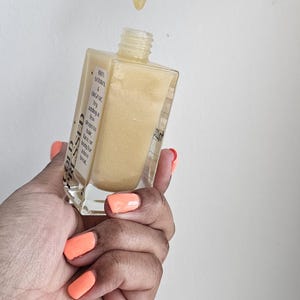 May include: A clear, square glass bottle filled with a golden liquid, with a dropper dispensing a drop. The bottle has a white label with text. The person's nails are painted orange.