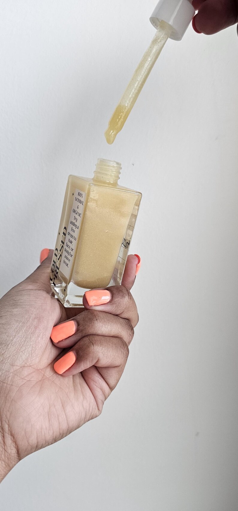 May include: A clear, square glass bottle filled with a golden liquid, with a dropper dispensing a drop. The bottle has a white label with text. The person's nails are painted orange.