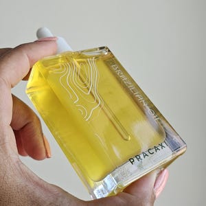 May include: A clear, rectangular bottle filled with yellow liquid, labeled "BRAZILIAN OIL" and "PRACAXI." The bottle has a white dropper and a white graphic design. The bottle is held in a hand with painted nails.