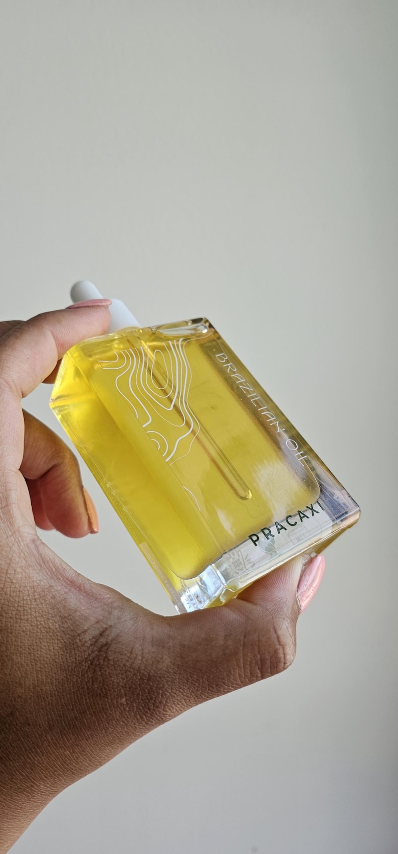 May include: A clear, rectangular bottle filled with yellow liquid, labeled "BRAZILIAN OIL" and "PRACAXI." The bottle has a white dropper and a white graphic design. The bottle is held in a hand with painted nails.
