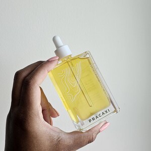 May include: A clear, rectangular bottle of Pracaxi oil with a white dropper top. The yellow liquid inside has a white topographical design. The bottle is held against a white background, with the word "PRACAXI" printed on the front.