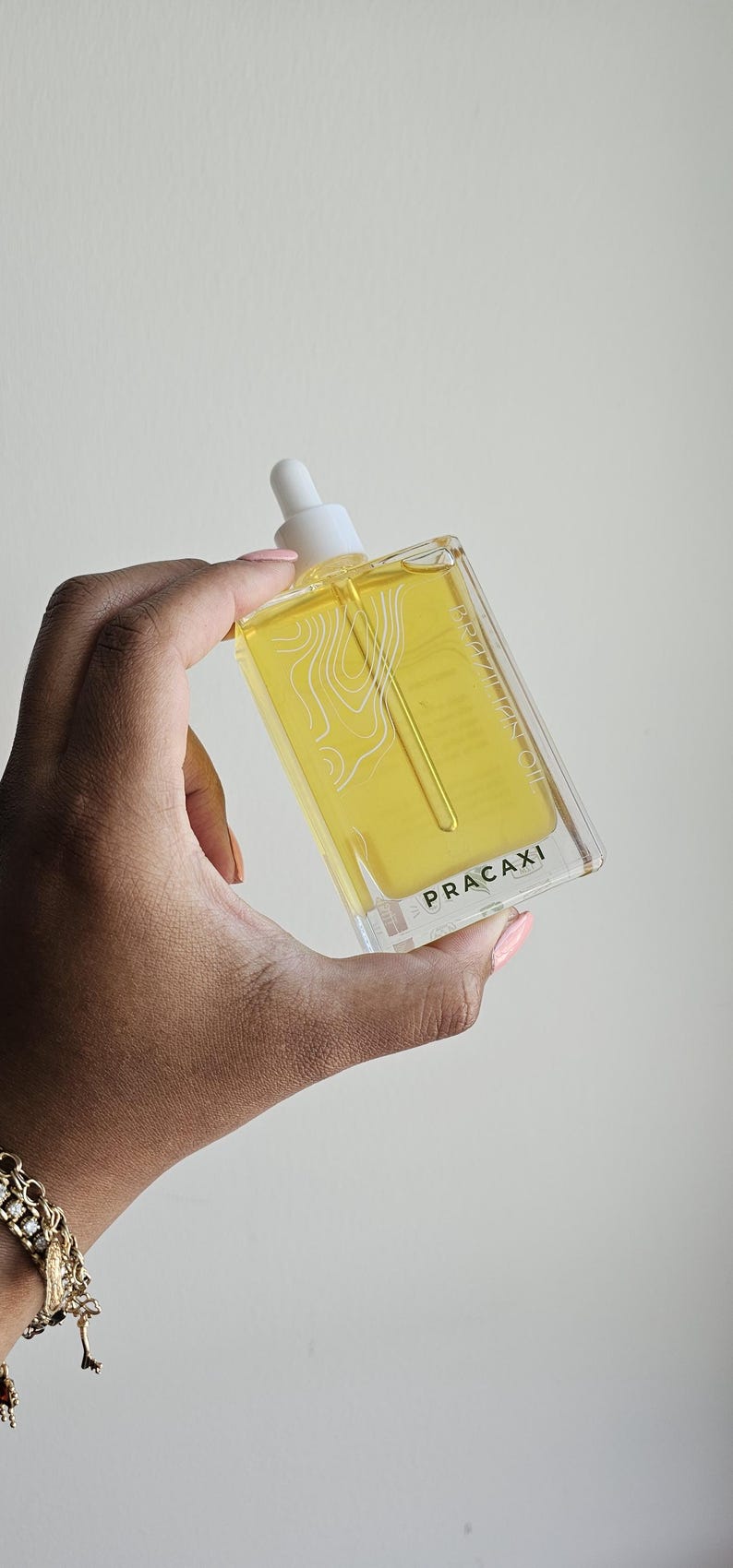 May include: A rectangular glass bottle filled with yellow liquid, held in a hand. The bottle has a white dropper top and a white label with the word "PRACAXI" and a white topographic design. The bottle is approximately 4 inches tall.