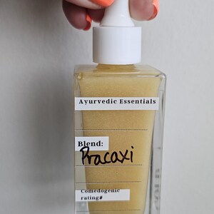 May include: A clear, square glass bottle with a white dropper top, filled with a golden liquid. The label reads "Ayurvedic Essentials" and "Blend: Pracaxi." The bottle is held by a hand with orange nail polish.