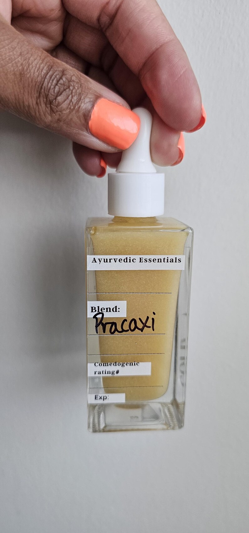 May include: A clear, square glass bottle with a white dropper top, filled with a golden liquid. The label reads "Ayurvedic Essentials" and "Blend: Pracaxi." The bottle is held by a hand with orange nail polish.