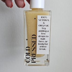 May include: A clear, rectangular bottle of cold-pressed oil with a white cap. The bottle contains a yellow liquid and has a label with text that reads "100% Natural & Organic". The label also suggests using the oil on hair, face, or body.