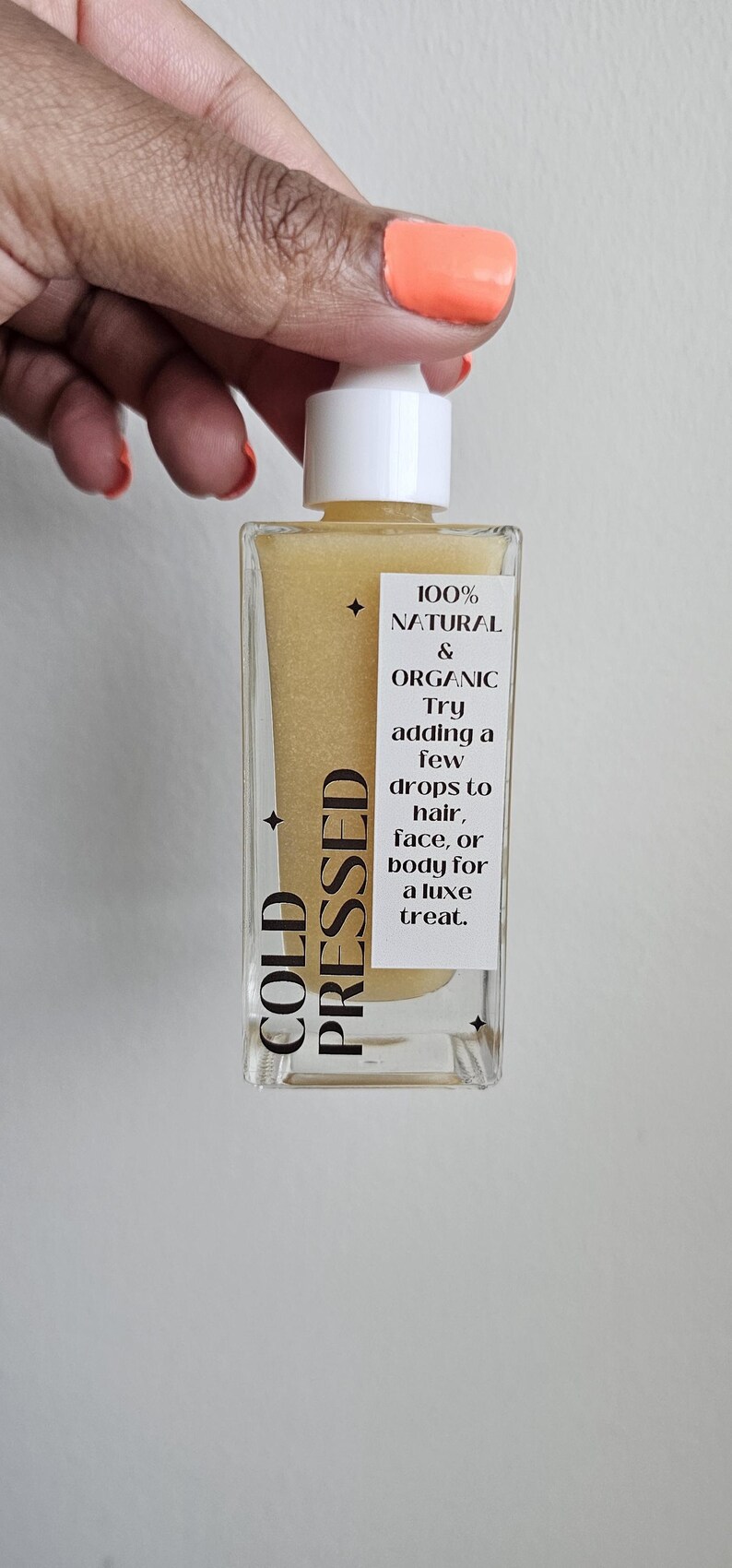 May include: A clear, rectangular bottle of cold-pressed oil with a white cap. The bottle contains a yellow liquid and has a label with text that reads "100% Natural & Organic". The label also suggests using the oil on hair, face, or body.