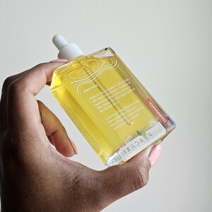 May include: A clear, rectangular bottle of hair and scalp oil is held in a hand. The bottle has a white dropper and contains a yellow liquid. Text on the bottle includes directions and product information. The brand name is visible.