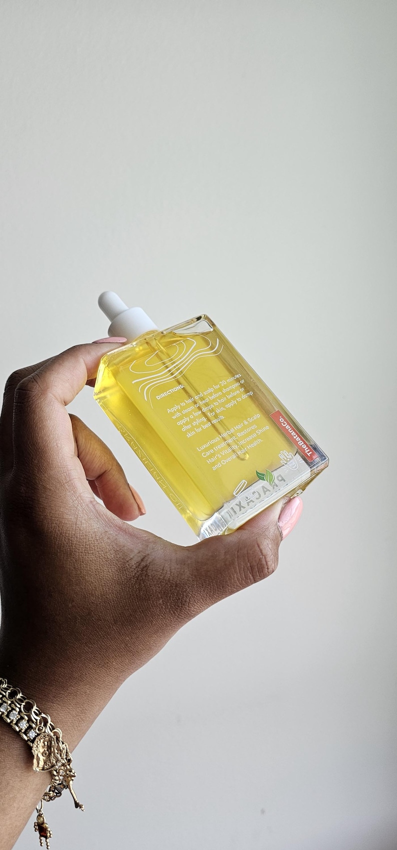 May include: A clear, rectangular bottle of hair and scalp oil is held in a hand. The bottle has a white dropper and contains a yellow liquid. Text on the bottle includes directions and product information. The brand name is visible.