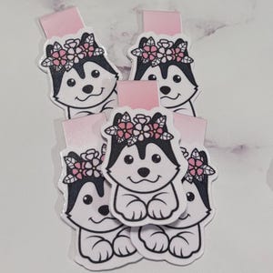 May include: Set of five pink and white magnetic bookmarks featuring a cartoon husky dog wearing a flower crown.