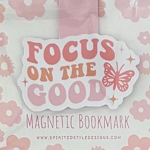 May include: Pink and orange magnetic bookmark with the text "Focus on the good" and a butterfly design. The bookmark also includes the text "Magnetic Bookmark" and a website address.