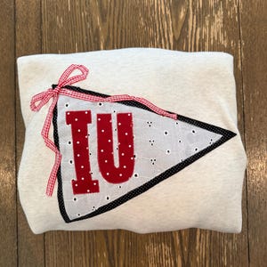 May include: A light gray sweatshirt with a red and white pennant design. The pennant features the letters "IU" in red, with a white dotted fabric background and a black border. A red and white gingham ribbon is tied at the top.