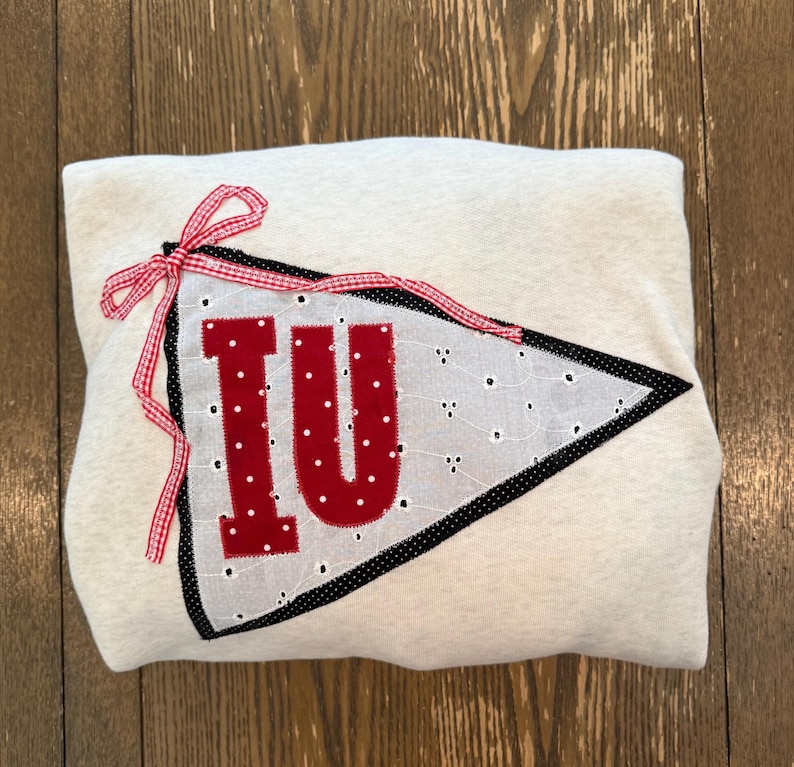 May include: A light gray sweatshirt with a red and white pennant design. The pennant features the letters "IU" in red, with a white dotted fabric background and a black border. A red and white gingham ribbon is tied at the top.