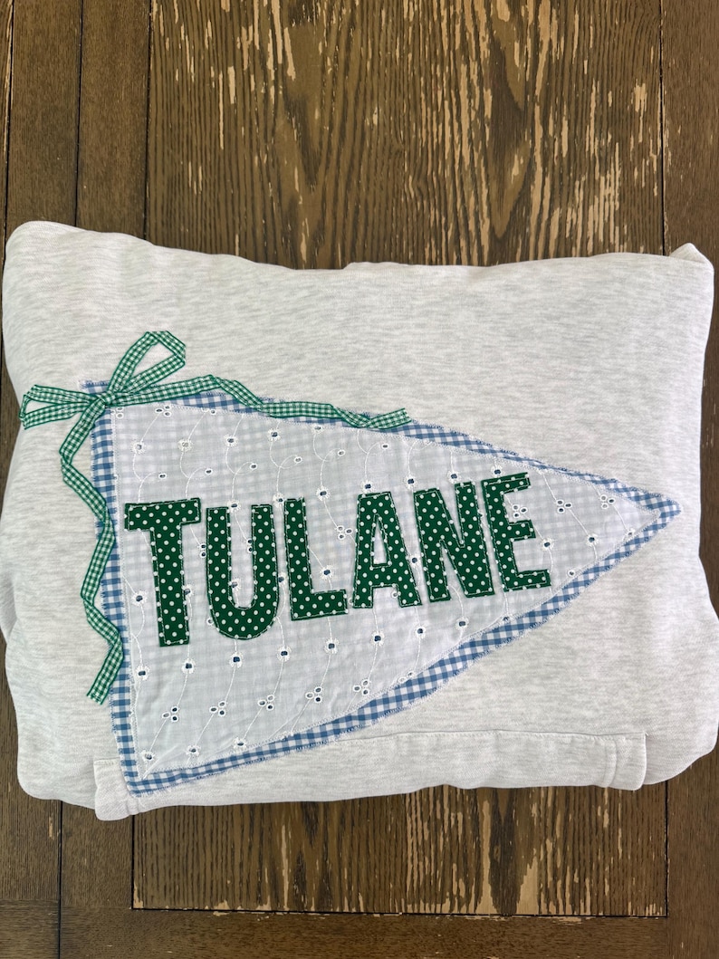 May include: A light gray hooded sweatshirt with a pennant-shaped patch. The patch is white with a blue gingham border and the word "TULANE" in green polka dot letters. A green gingham ribbon is tied at the top.
