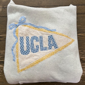 May include: A light gray pillow with a pennant design. The pennant is white with blue gingham letters spelling "UCLA". It has a yellow border and is tied with a blue gingham ribbon.