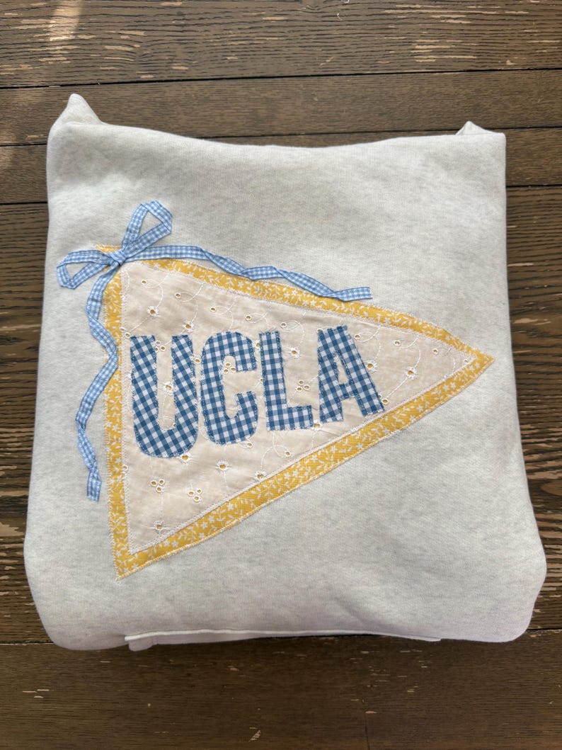 May include: A light gray pillow with a pennant design. The pennant is white with blue gingham letters spelling "UCLA". It has a yellow border and is tied with a blue gingham ribbon.