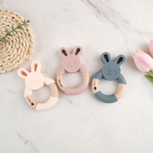 May include: Three wooden teething rings with silicone bunny heads. The rings are pink, blue, and light pink. Each ring has a name engraved on it: "Jack", "George", and "John".
