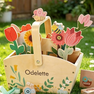 Pode incluir: Uma cesta de brinquedo de madeira cheia de flores de madeira coloridas, uma borboleta e uma abelha. A cesta tem uma alça e é decorada com desenhos florais e o nome "Odelette". A cena é ambientada em um gramado verde.