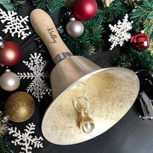 May include: A gold-coloured jingle bell with a wooden handle. The handle is engraved with the name "Kathy" and the date "02.23.2023".