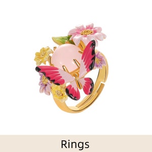 May include: A gold ring with a pink butterfly and floral design. The butterfly has pink and black wings and a pink body. The flowers are yellow, pink, and purple.