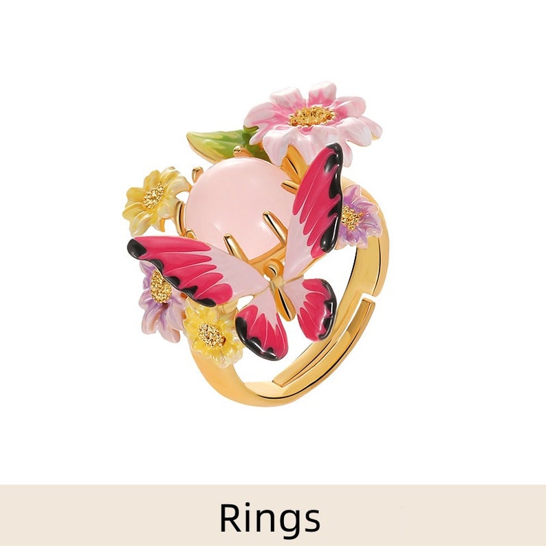 May include: A gold ring with a pink butterfly and floral design. The butterfly has pink and black wings and a pink body. The flowers are yellow, pink, and purple.
