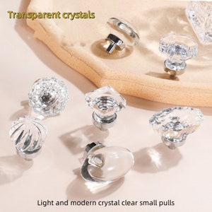 Könnte beinhalten: Sortiment an klaren Kristall-Schrankknöpfen mit silbernen Metallsockeln. Die Knöpfe haben verschiedene Formen, darunter rund, quadratisch, oval und achteckig. Transparente Kristalle.