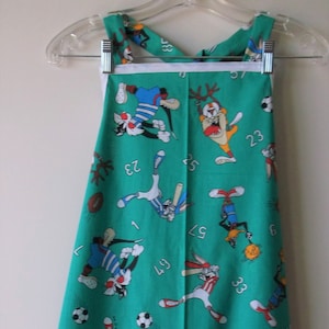 May include: A green apron with a white trim and a cartoon character print. The characters are playing sports and the numbers 1, 3, 5, 7, 9, 10, 23, 33, 49, 57, 67, and 98 are printed on the apron.