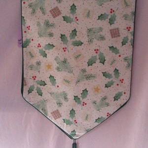 May include: A green and white table runner with a holly and pine needle pattern. The runner has a green tassel and a scalloped edge.