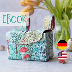 Op de afbeelding: Een groene en witte stoffen tas met een rode handgreep en een bosprint. De tas heeft een klep met een drukknopsluiting en een klein vakje aan de voorkant. De tekst "EBOOK" is in turquoise op een witte achtergrond geschreven. De tas ligt op een wit oppervlak met een Duitse en Amerikaanse vlag op de achtergrond.