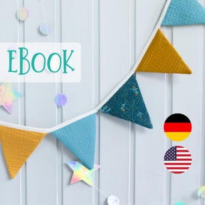 Könnte beinhalten: Eine blaue und gelbe Stoffgirlande mit weißem Besatz hängt an einer weißen Holzwand. Die Girlande ist mit einem Blumenmuster verziert. Das Wort "EBOOK" ist in grünen Buchstaben auf einem weißen Rechteck geschrieben. Das Bild zeigt auch zwei Flaggen: eine deutsche Flagge und eine amerikanische Flagge.