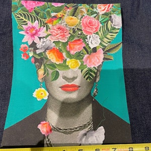 May include: A fabric patch featuring a portrait of a woman with a floral headdress. The image has a teal background and the woman is wearing a black jacket, silver necklace, and earrings. The woman's lips are red, and the headdress is composed of various colorful flowers.